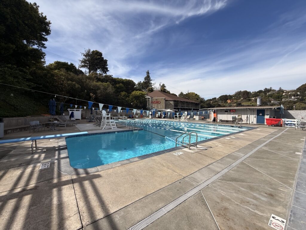 Outdoor swimming pool with lane dividers, lifeguard chairs, and poolside seating on a sunny day; buildings and trees are visible in the background.