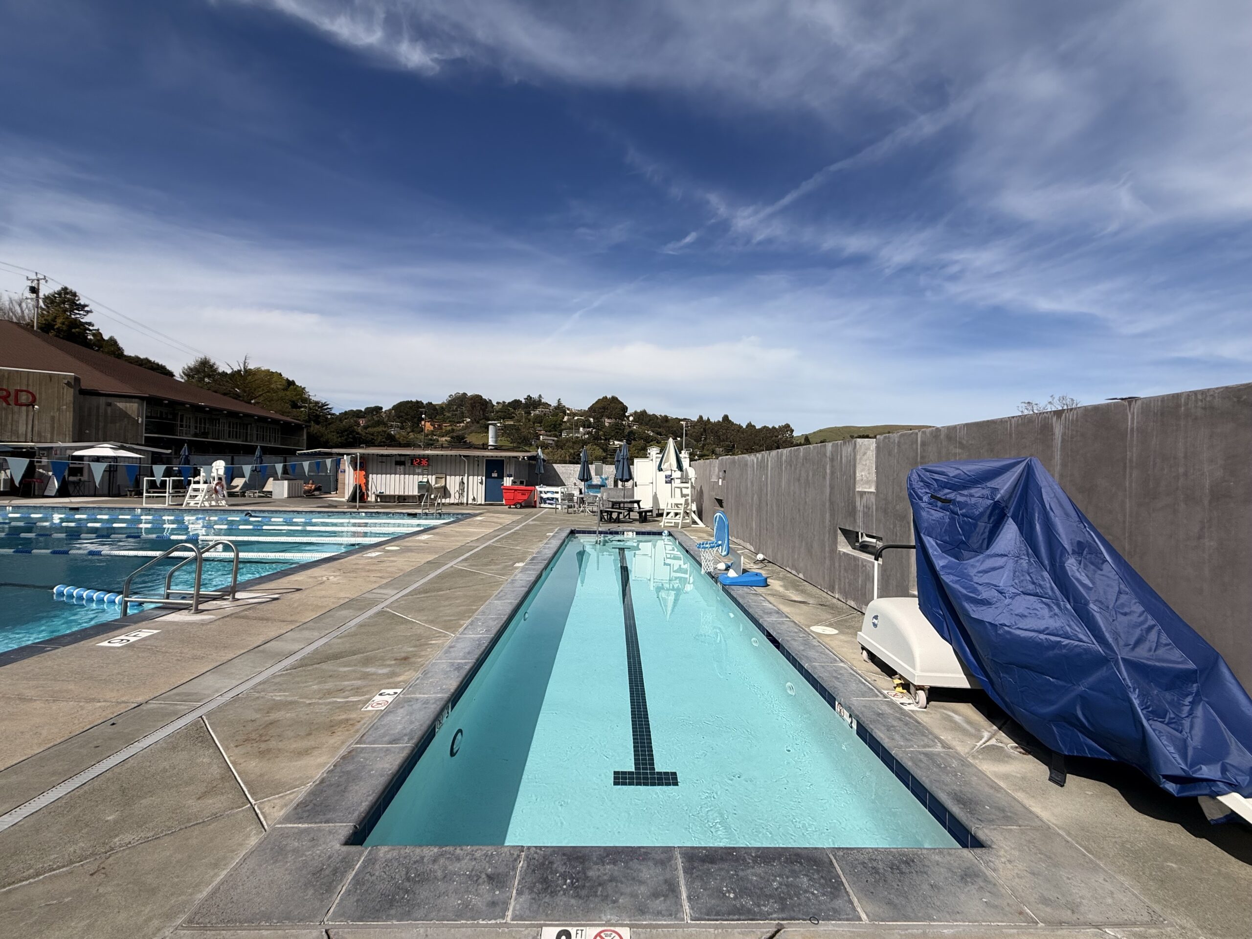 A narrow outdoor pool with clear water and a black center line is next to a larger lap pool; pool equipment is covered by a blue tarp on the right.