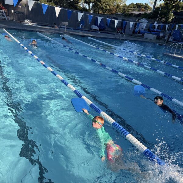 Children swim in outdoor pool lanes using kickboards, wearing goggles and swim caps, with lane dividers and blue flags overhead.