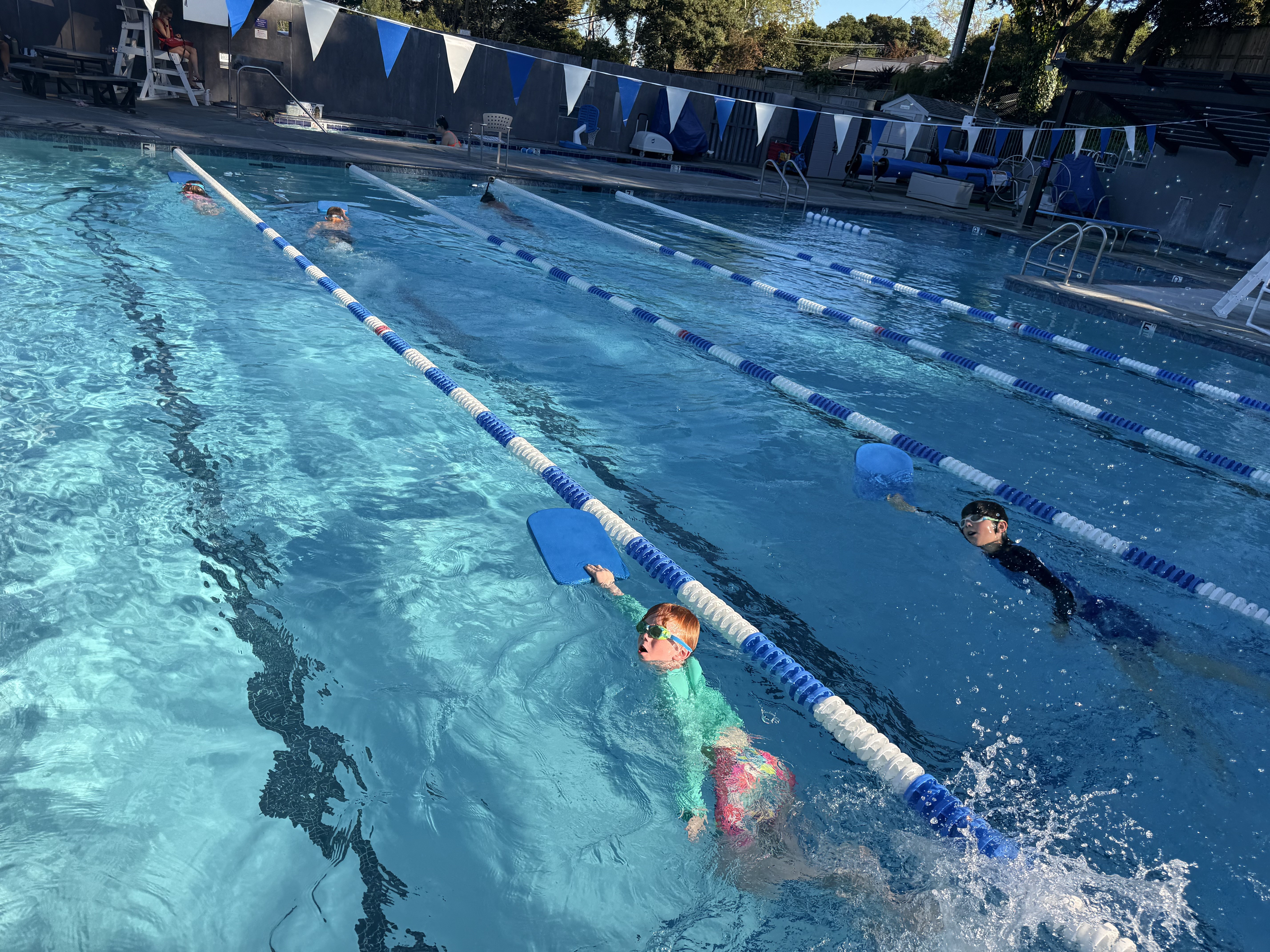 Children swim in outdoor pool lanes using kickboards, wearing goggles and swim caps, with lane dividers and blue flags overhead.