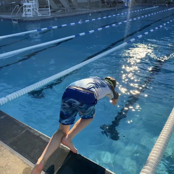 A person in swim trunks and a swim cap dives into a sunlit outdoor swimming pool, with empty lanes visible in the background.