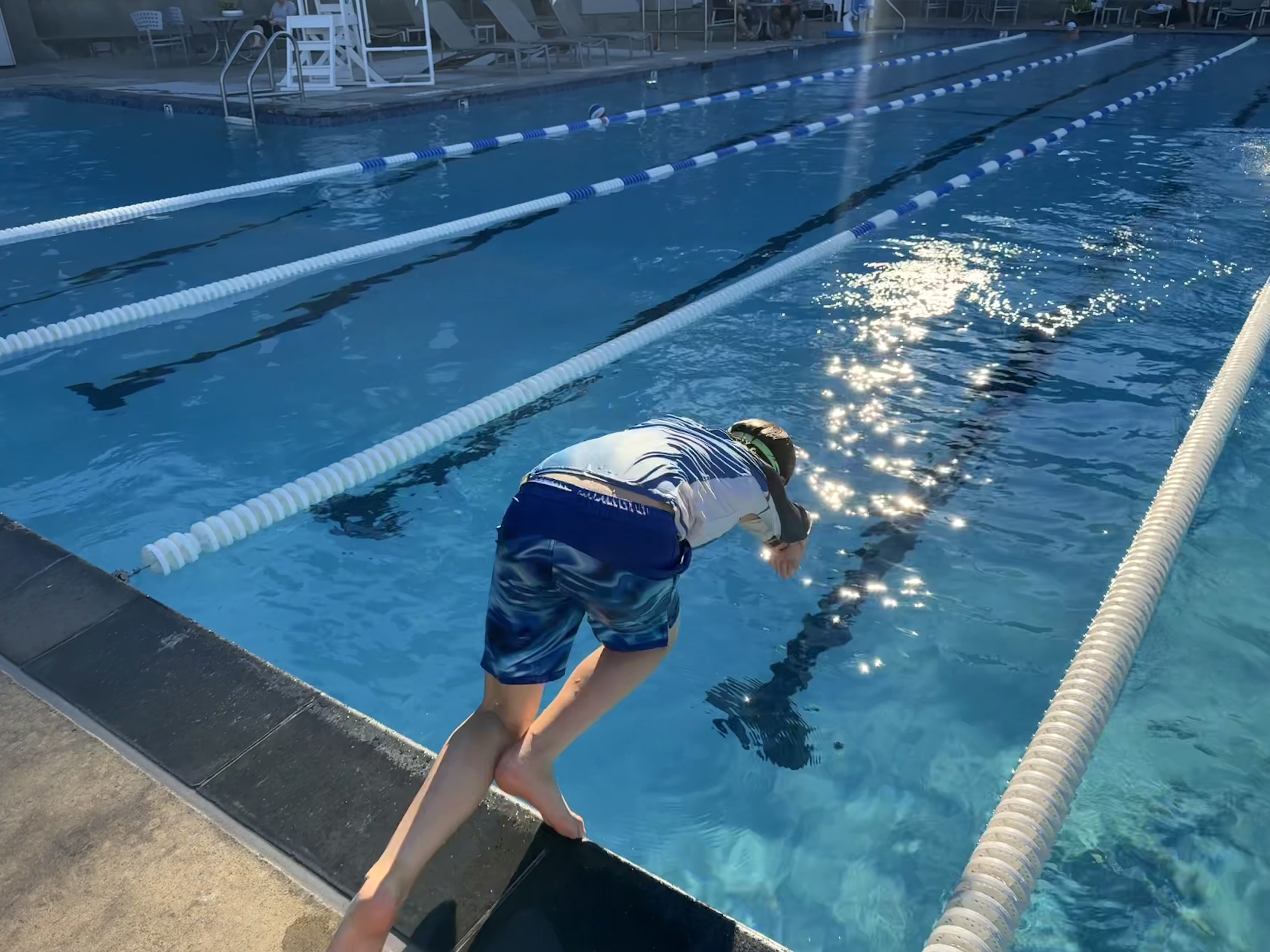 A person in swim trunks and a swim cap dives into a sunlit outdoor swimming pool, with empty lanes visible in the background.