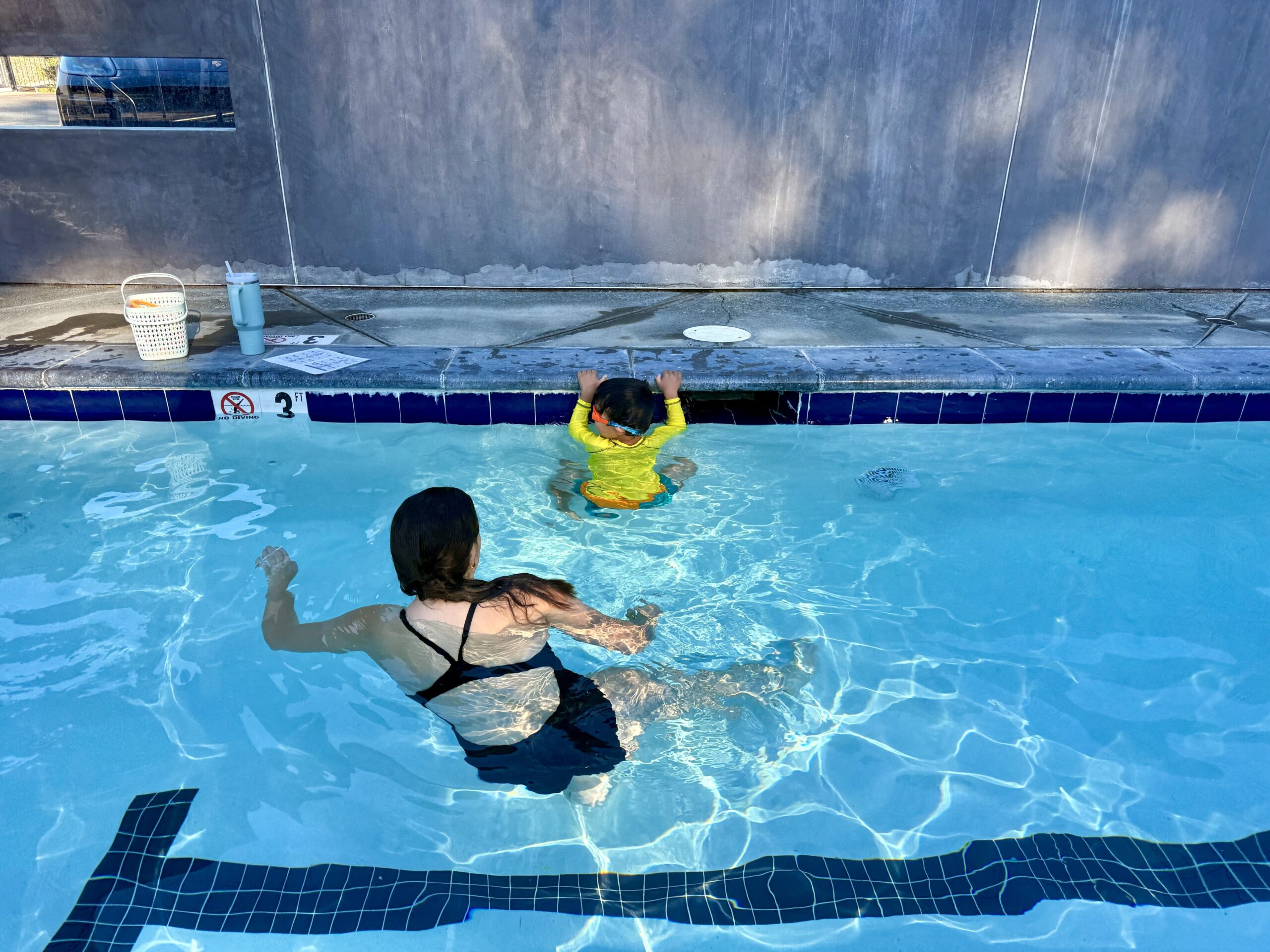 An adult and a child are in a swimming pool; the adult faces the child, who is holding onto the pool edge near a wall. A white basket and bottle sit on the poolside.