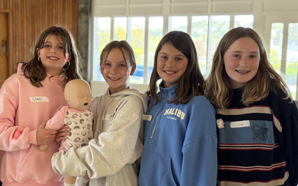 Four girls stand indoors, smiling at the camera; one girl holds a baby doll. Each girl wears a hoodie or sweater and has a name tag. Bright windows are visible in the background.