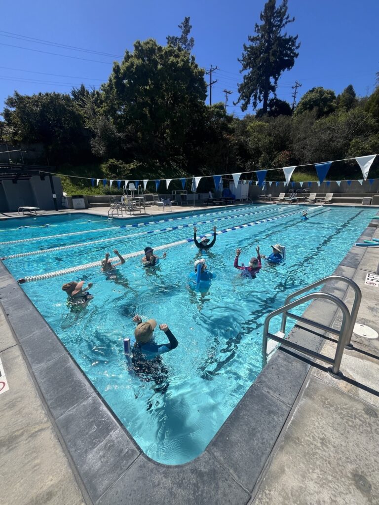 A group of people participate in a water exercise class in an outdoor swimming pool on a sunny day.