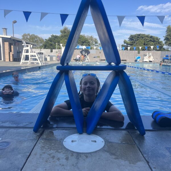 A child in a swimming pool poses behind a structure made of blue pool noodles stacked in a triangular shape at the pool edge. Other swimmers and poolside equipment are visible in the background.