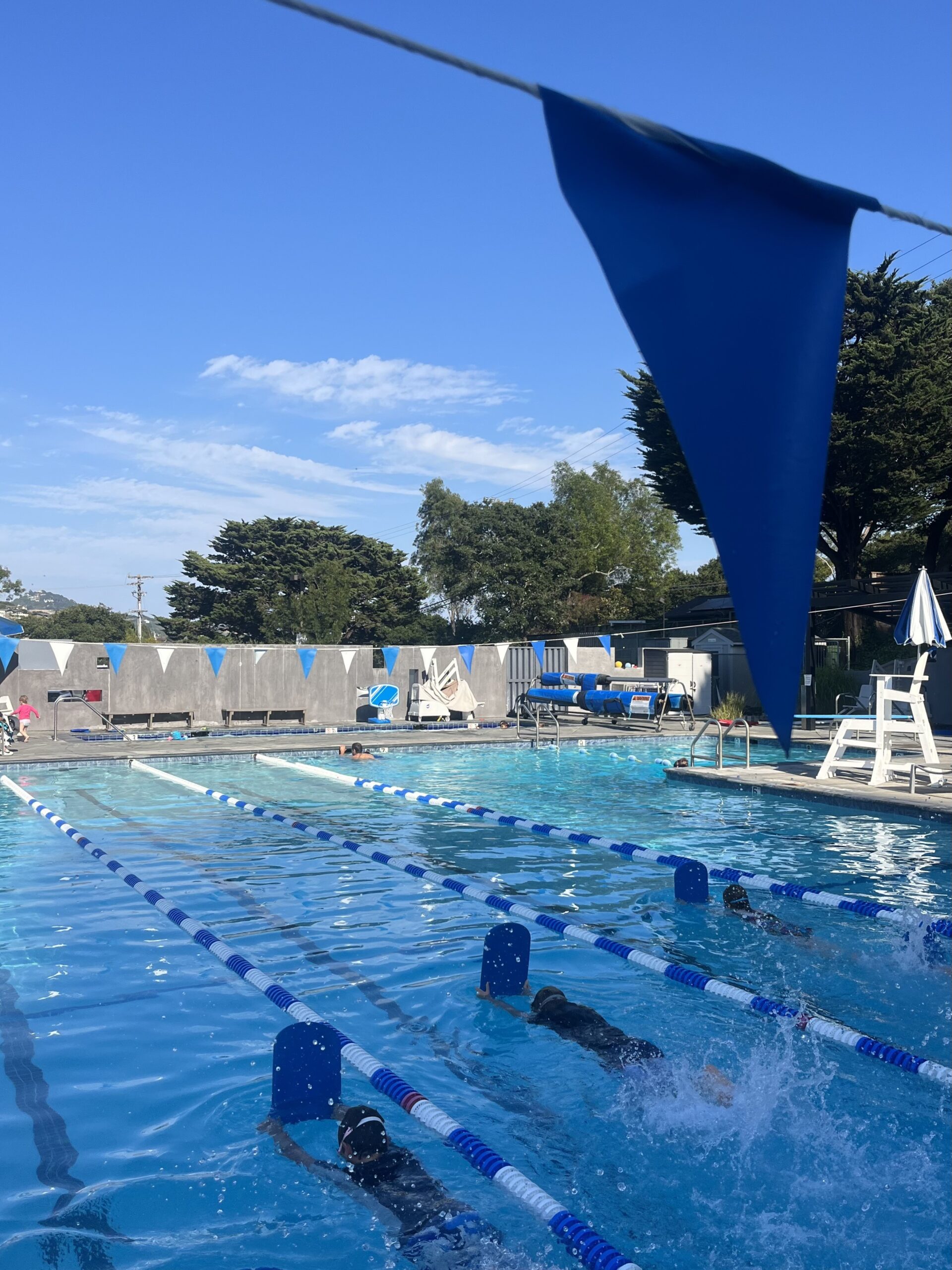 Several swimmers practice strokes with kickboards in an outdoor pool under a clear blue sky, with lane dividers and a blue pennant in the foreground.