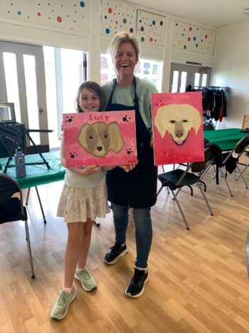 An adult and a child stand indoors holding dog paintings, smiling. The child’s painting says "Lucy." Tables and chairs are arranged in the background.