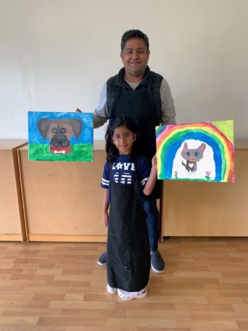 A man and a young girl stand indoors, each holding a colorful painting featuring a dog. The girl stands in front wearing a black apron.