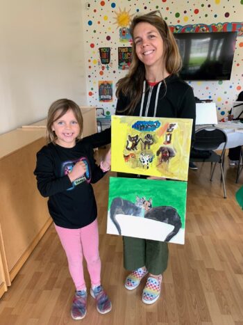A woman and a young girl stand indoors, smiling and holding two colorful cat paintings. The background has polka dot decorations and a large TV.