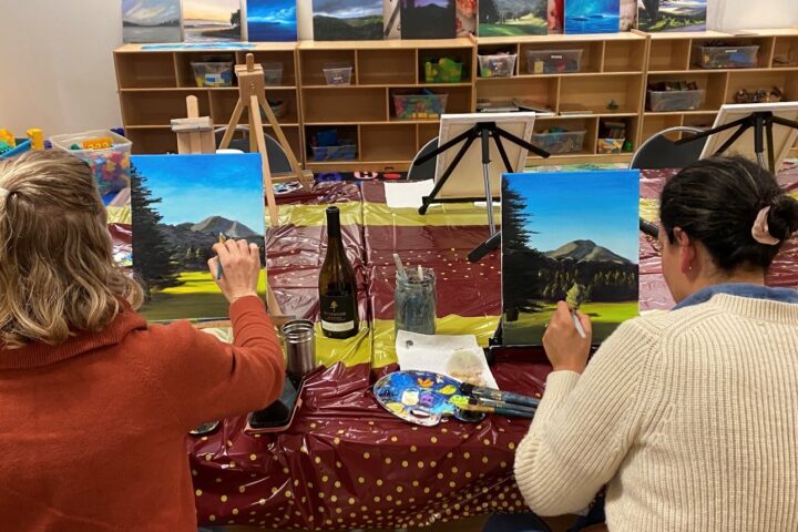 Two people sit at a table painting landscapes on canvases; art supplies, a wine bottle, and finished paintings are visible in the background.