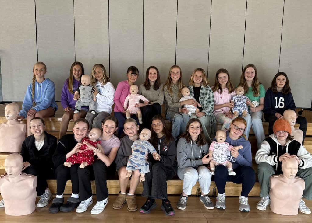 A group of teenagers sit on bleachers holding infant mannequins and CPR training dummies, posing for a photo in a gym or classroom setting.