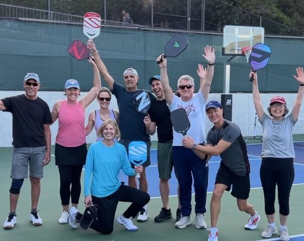A group of eleven people pose and smile with pickleball paddles on an outdoor court, some raising their arms in celebration.