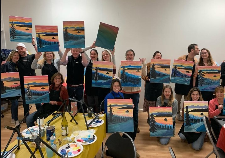 A group of people in a classroom display their finished sunset landscape paintings while posing for a group photo. Art supplies and palettes are visible on the tables.