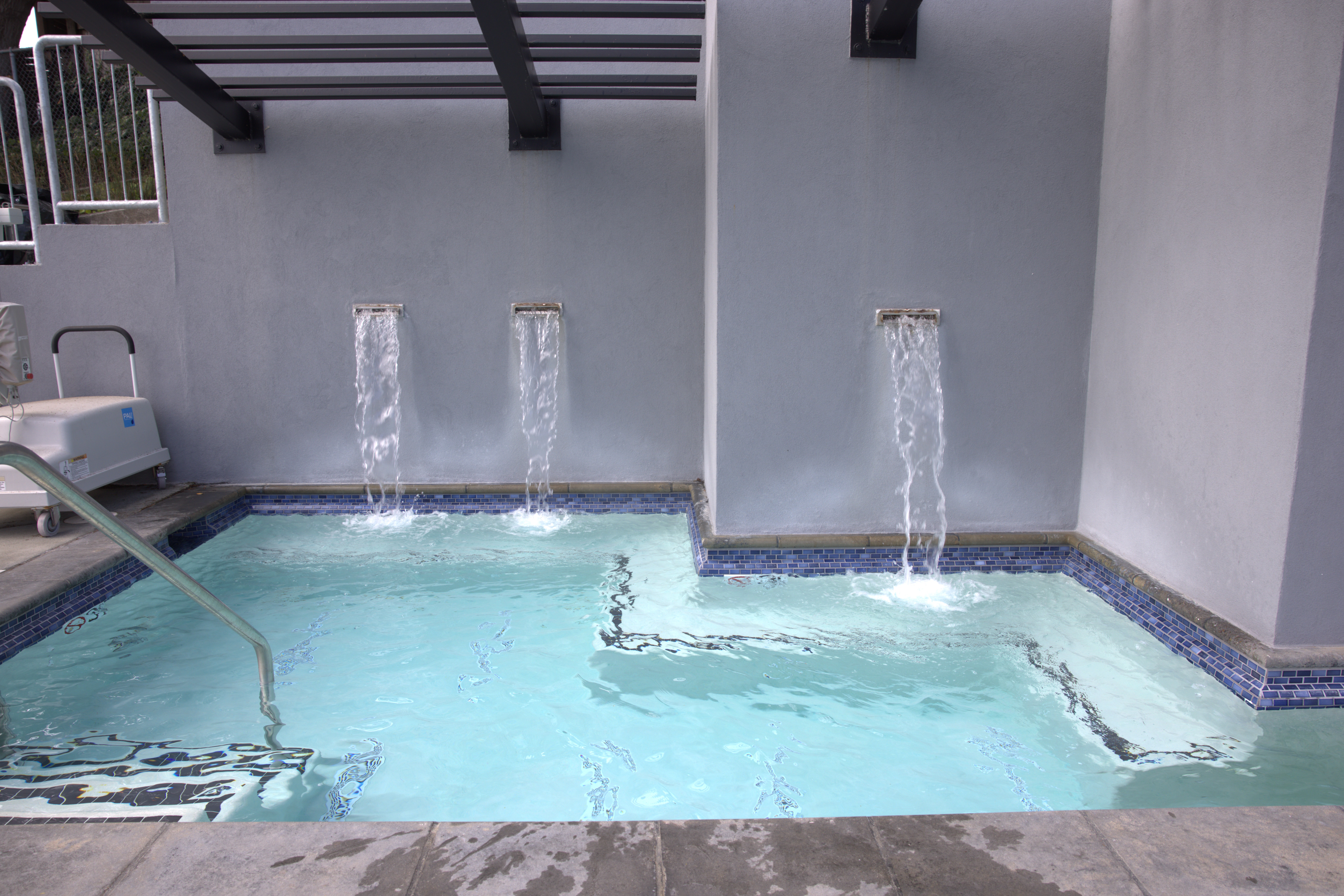 Small indoor pool with three water spouts pouring water into it; metal handrail on the left and blue tile accents along the pool edge.