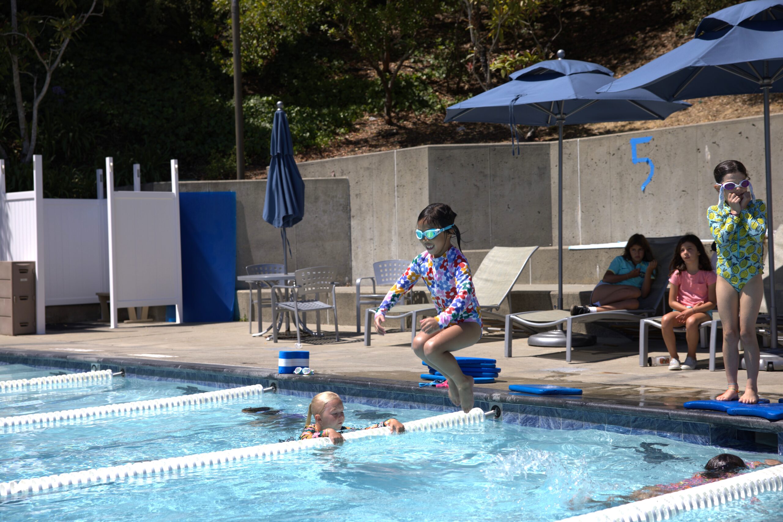 A child in swimwear and goggles jumps into a swimming pool while another child swims nearby; three people watch from poolside lounge chairs under umbrellas.