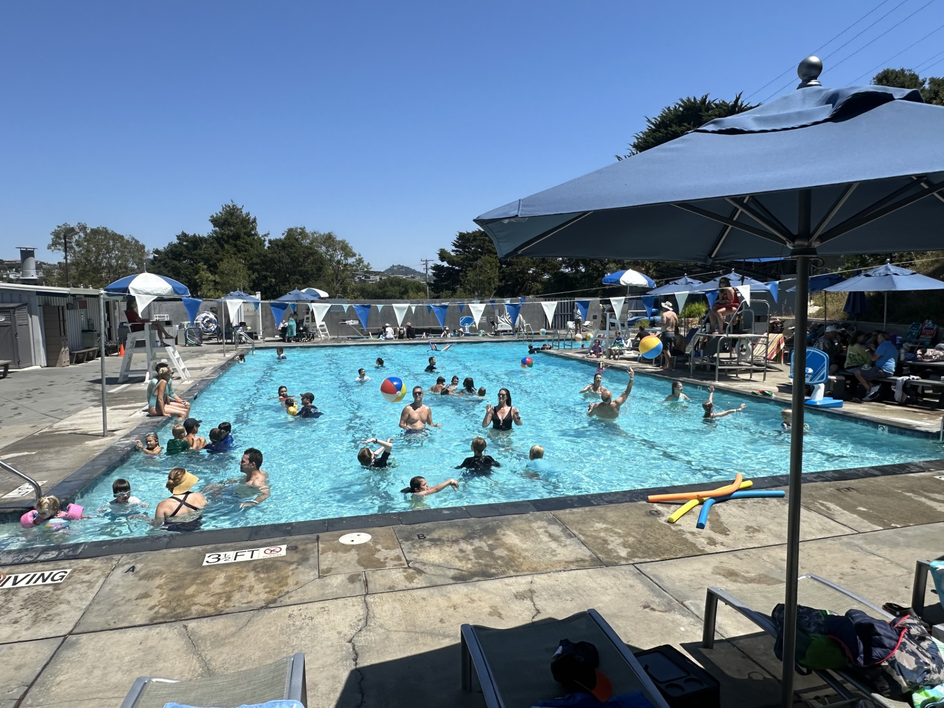 Outdoor swimming pool with many people swimming and playing with beach balls; umbrellas and lounge chairs line the poolside on a sunny day.