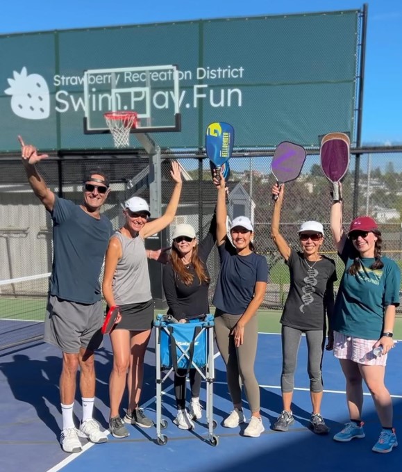 Six adults stand and smile on an outdoor court, holding up pickleball paddles, with a sign reading "Swim. Play. Fun" in the background.