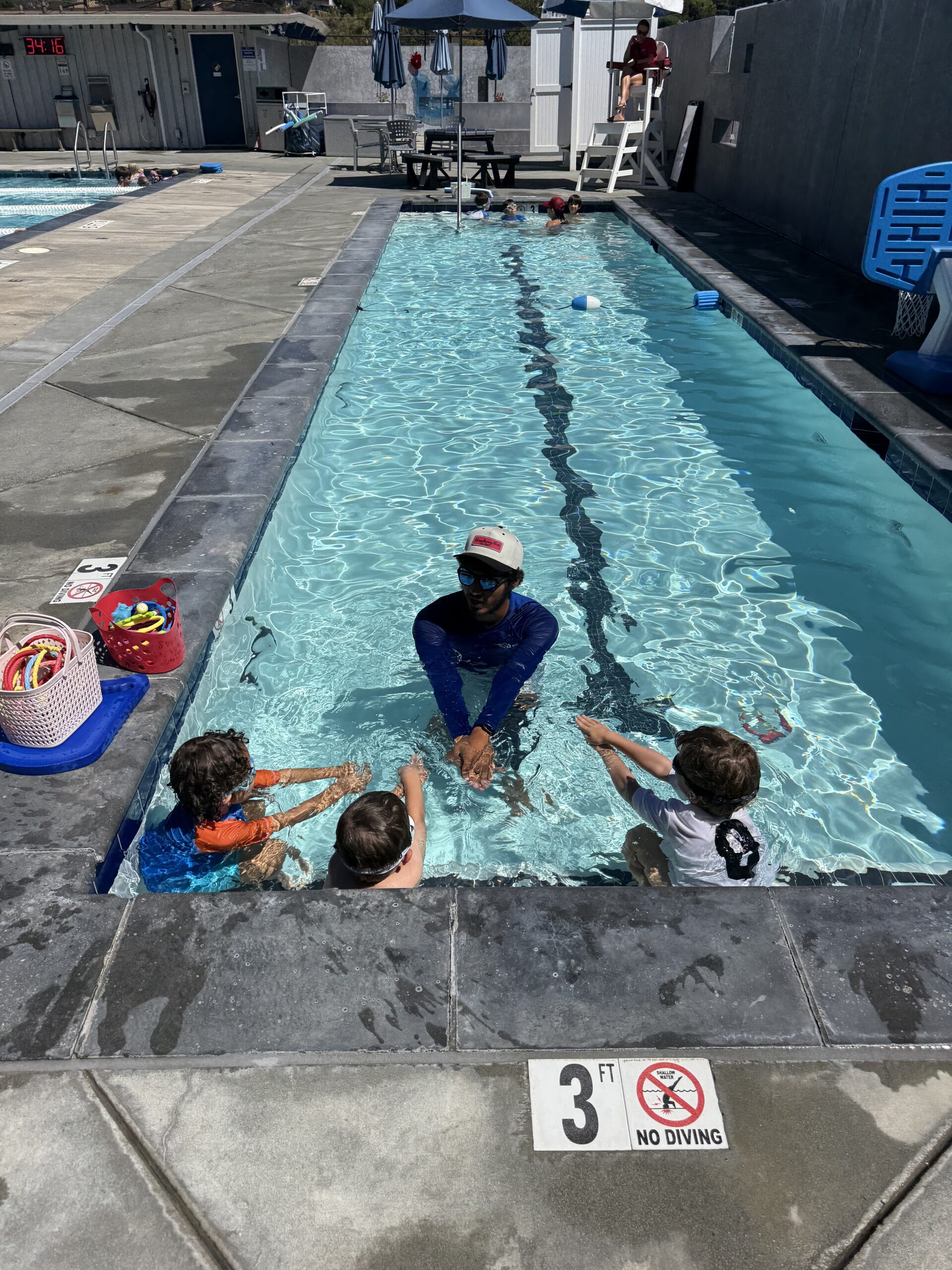 Three young children listen to an instructor in a shallow outdoor pool, with toys and bags nearby. A 