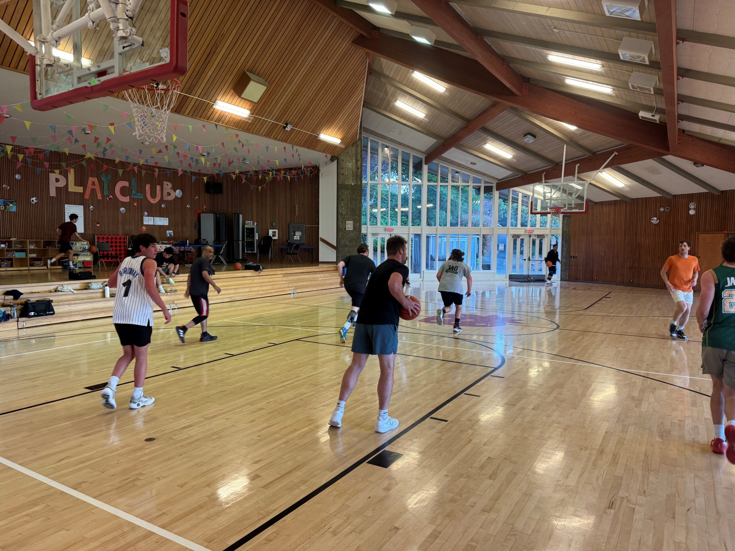 A group of people play basketball in a gymnasium with wooden floors and high ceilings. Benches and banners are visible in the background.