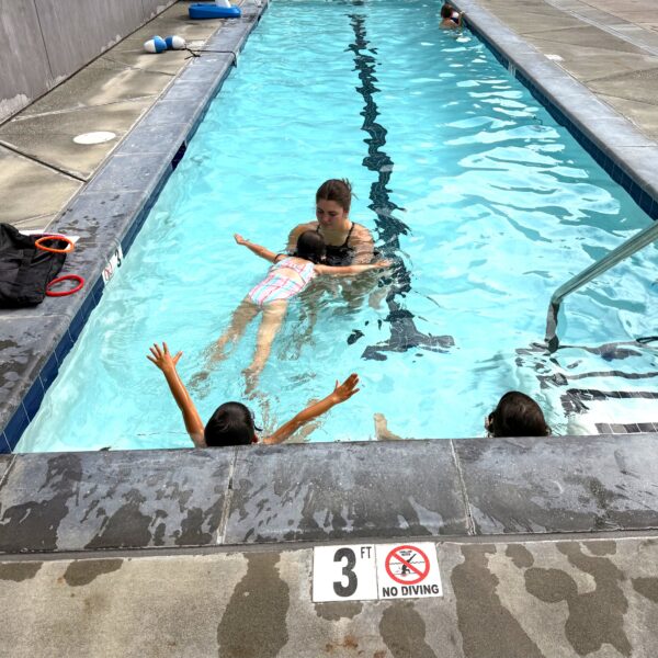 An adult teaches a young child to float on their back in a shallow swimming pool while two other children watch from the pool edge.