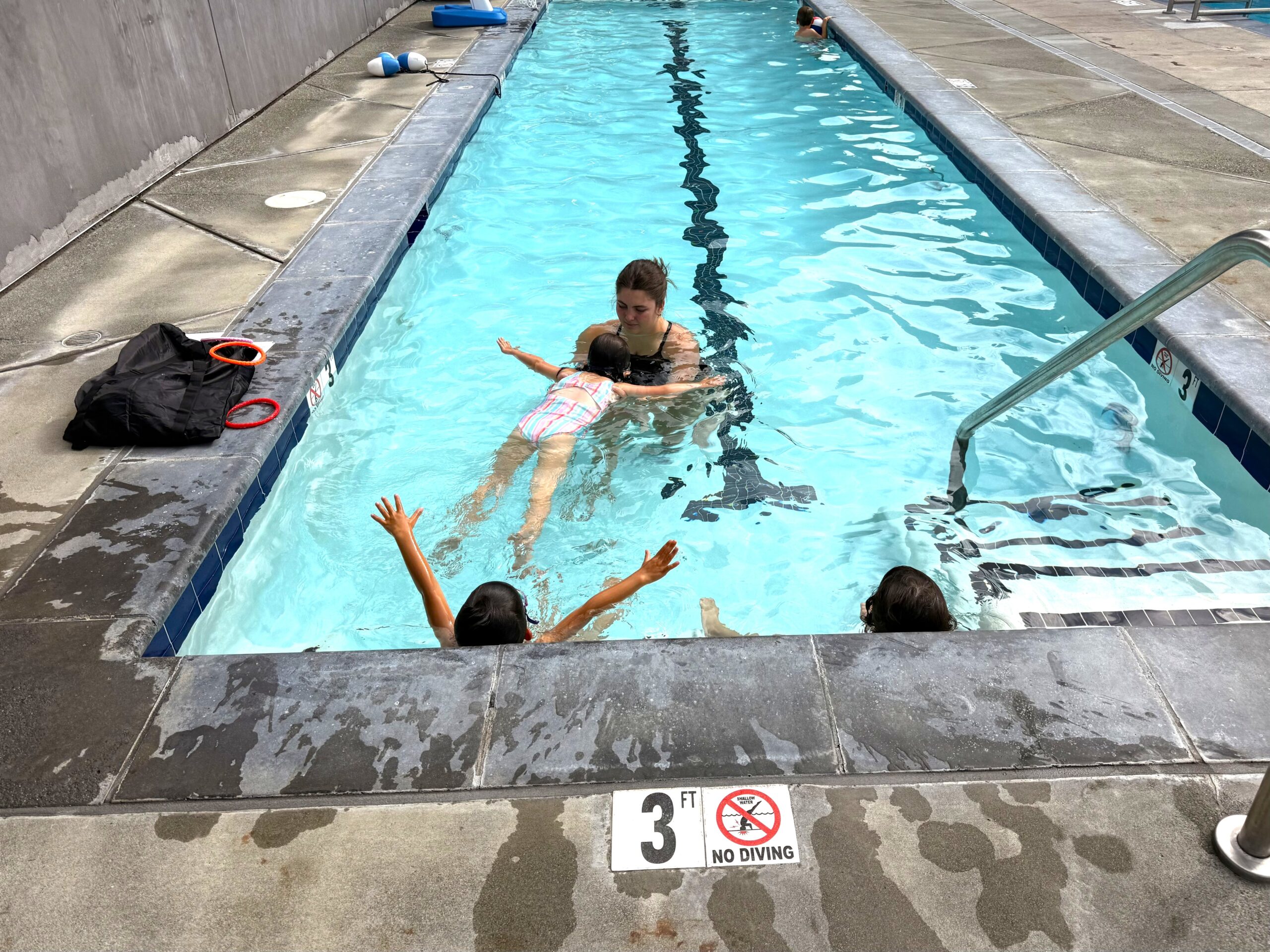 An adult teaches a young child to float on their back in a shallow swimming pool while two other children watch from the pool edge.