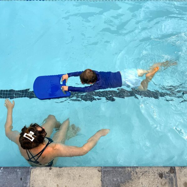 A child in swimwear uses a blue kickboard while a woman, possibly an instructor, guides them in a swimming pool near the edge.