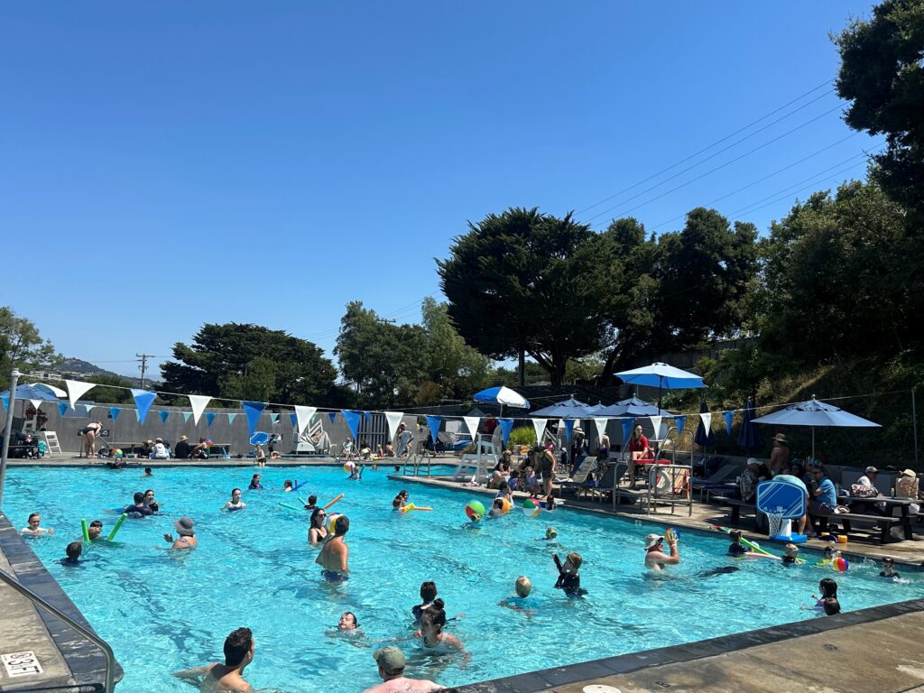 Outdoor public swimming pool with many people swimming and playing in the water; others sit under umbrellas on the poolside. It is a sunny day with clear blue sky.