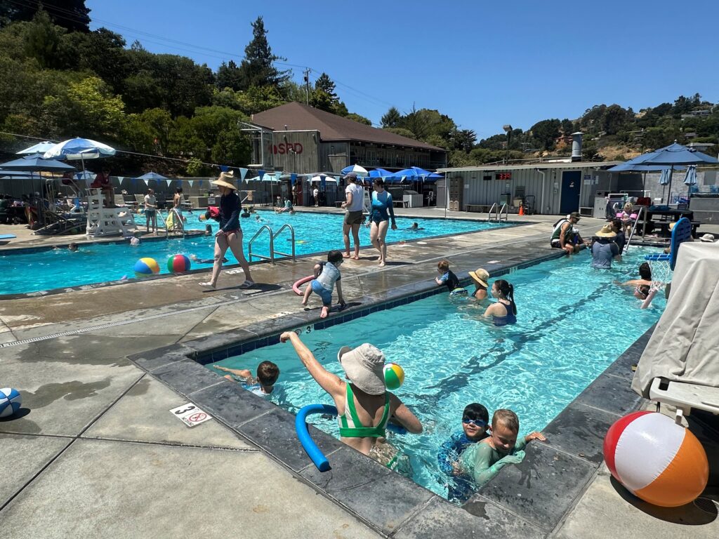 People, including children and adults, swim and play with pool toys in two outdoor swimming pools on a sunny day, with lounge chairs and umbrellas nearby.
