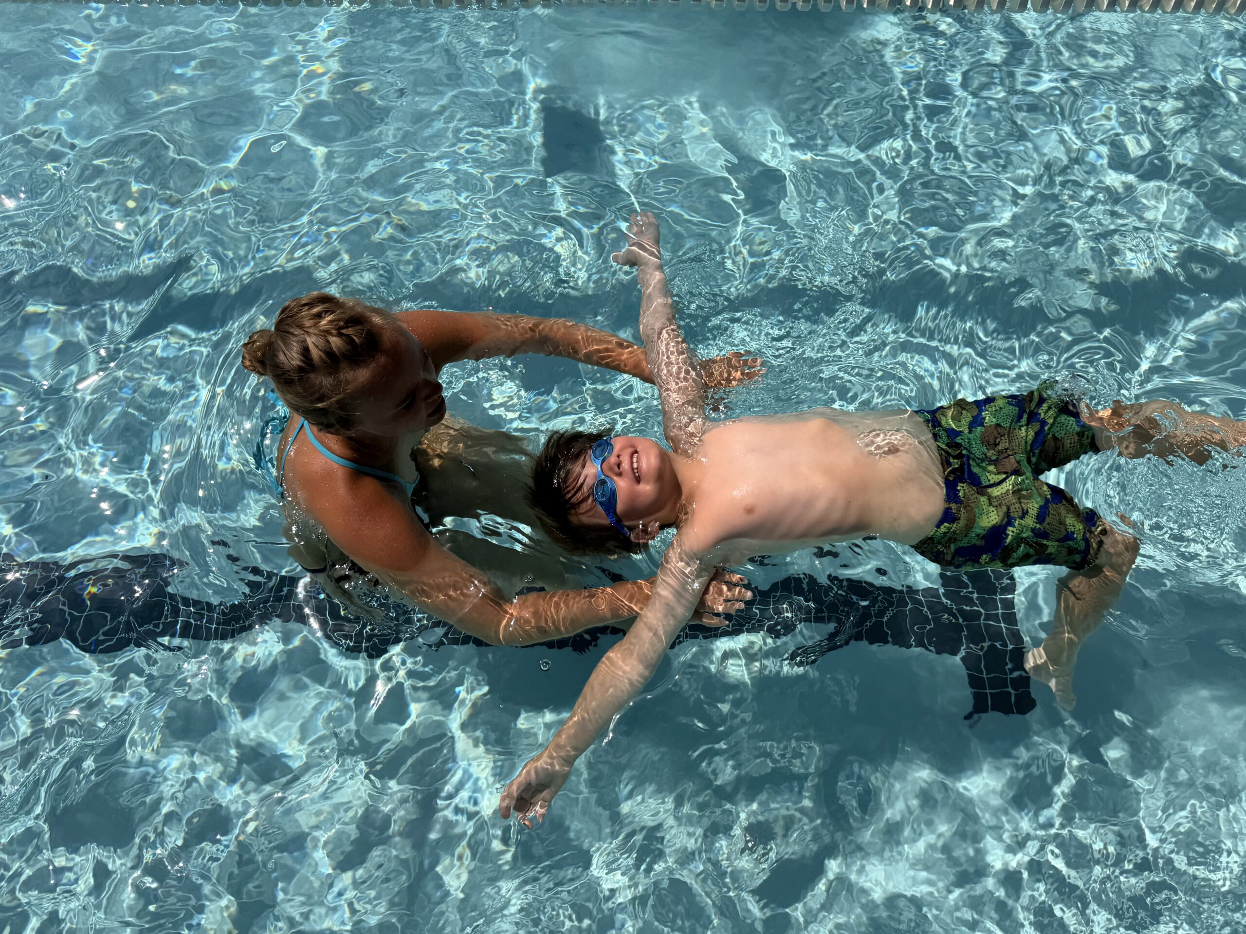 An adult helps a child float on his back in a swimming pool. The child wears goggles and swim trunks. Sunlight reflects on the water’s surface.