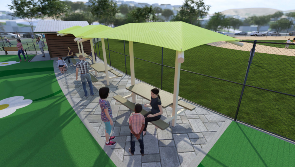People gather around picnic tables under green shade structures beside a fenced sports field on a sunny day; children and adults are present.