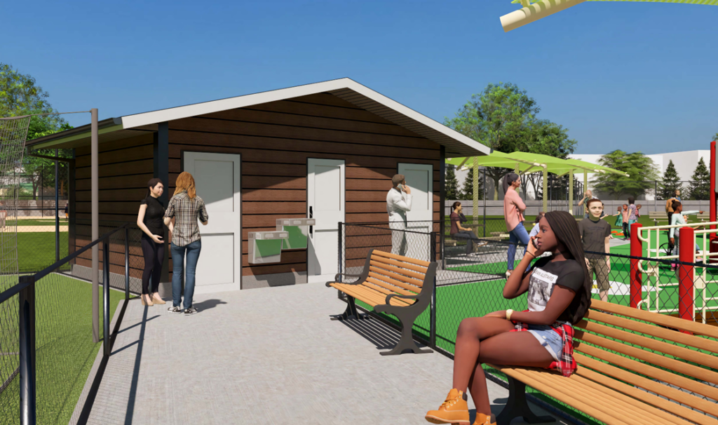People are gathered near a small building and benches in an outdoor park area with a playground and green space under a clear blue sky.