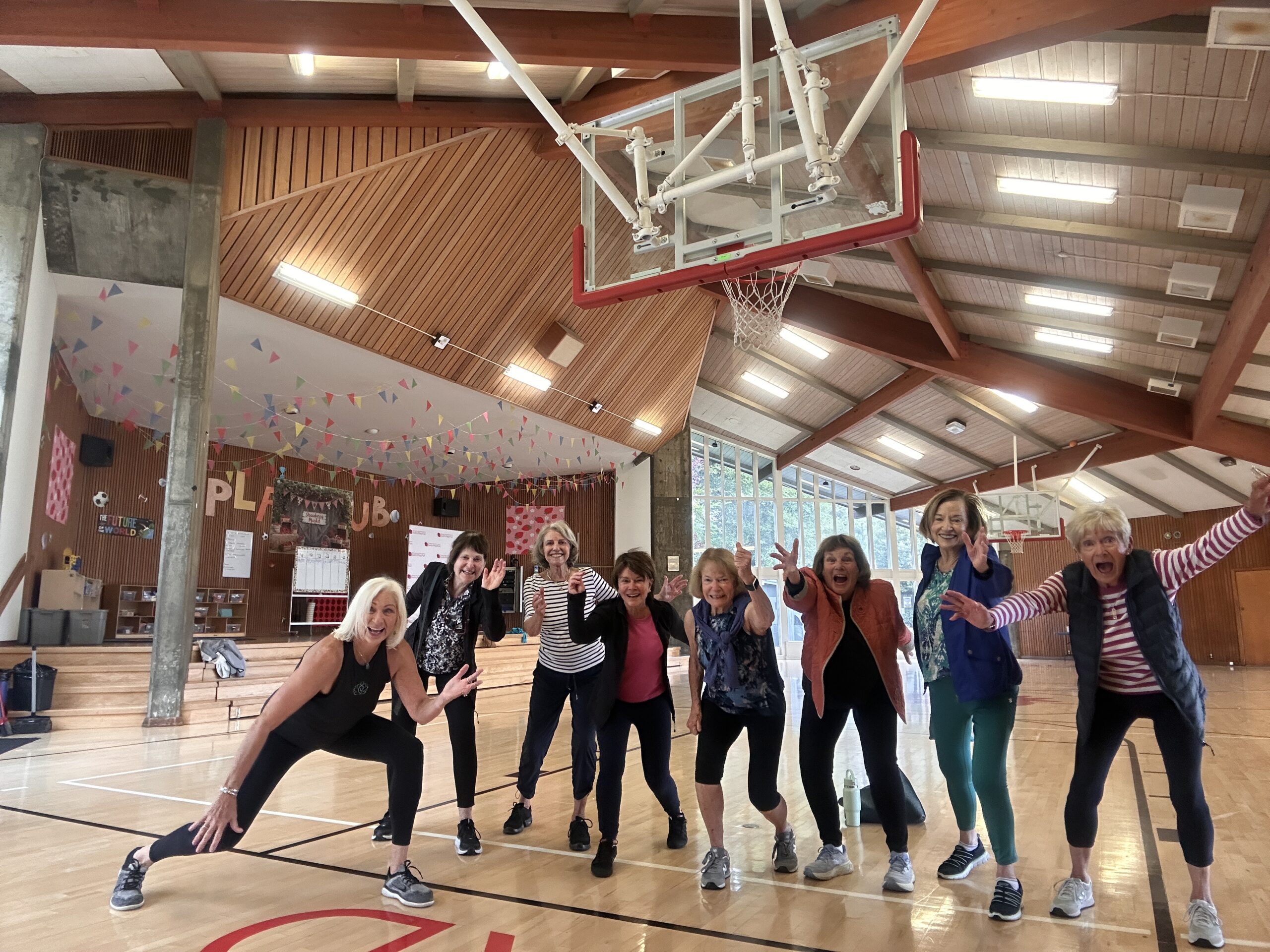 Nine women pose energetically under a basketball hoop in a gymnasium with wooden ceilings, large windows, and colorful pennant decorations.