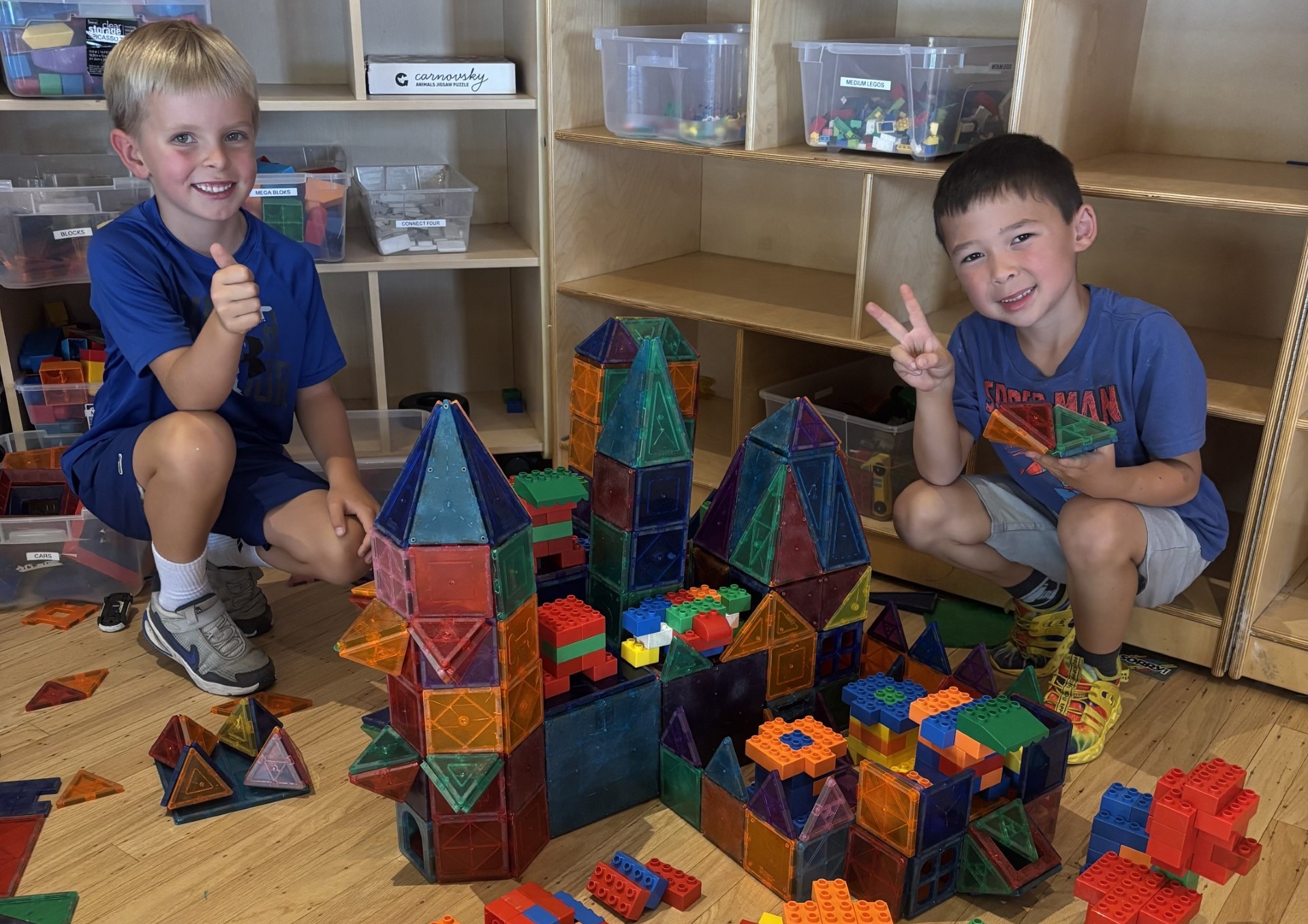 Two young boys smile and gesture with their hands while sitting beside a large, colorful structure built from magnetic tiles and plastic blocks in a classroom.